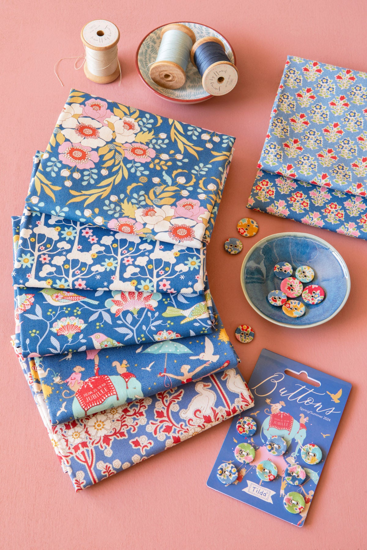 A styled flat-lay of sewing and fabric accessories on a pink background, including two stacks of blue patterned fabric, spools of thread, buttons in a small bowl, and a card of buttons.
Photo by Tildafabrics.com [additional_image_link]