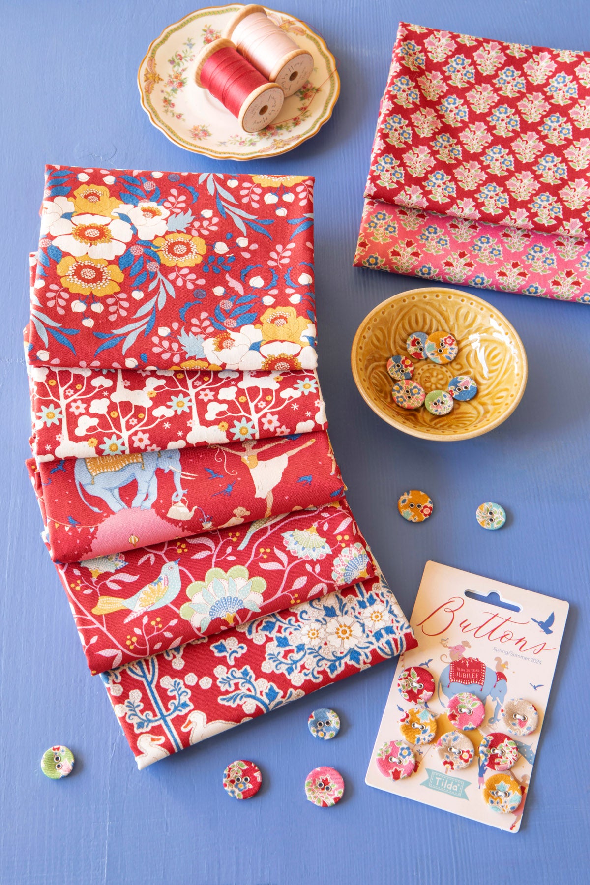 A styled flat-lay of fabric and buttons on a blue surface. Multiple folded and stacked pieces of red patterned fabric are arranged on the left and right, with floral and bird motifs.
Photo by Tildafabrics.com [additional_image_link]