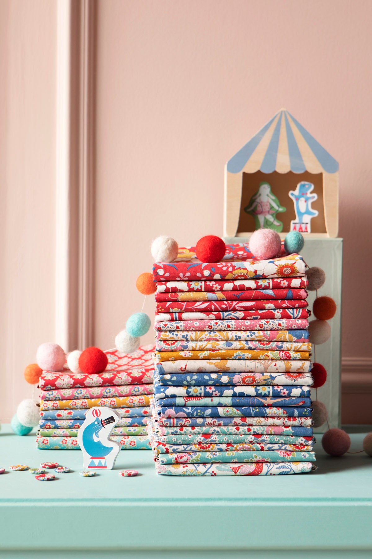 Stacks of colorful patterned fabric bundles arranged on a mint green surface against a dusty pink wall, with small decorative props including pom-poms, a small toy cabin, and paper cutouts.
Photo by Tildafabrics.com [additional_image_link]