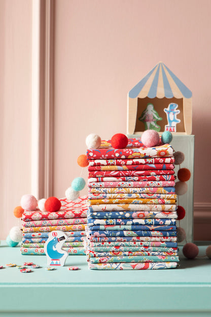 Stacks of colorful patterned fabric bundles arranged on a mint green surface against a dusty pink wall, with small decorative props including pom-poms, a small toy cabin, and paper cutouts.
Photo by Tildafabrics.com [additional_image_link]