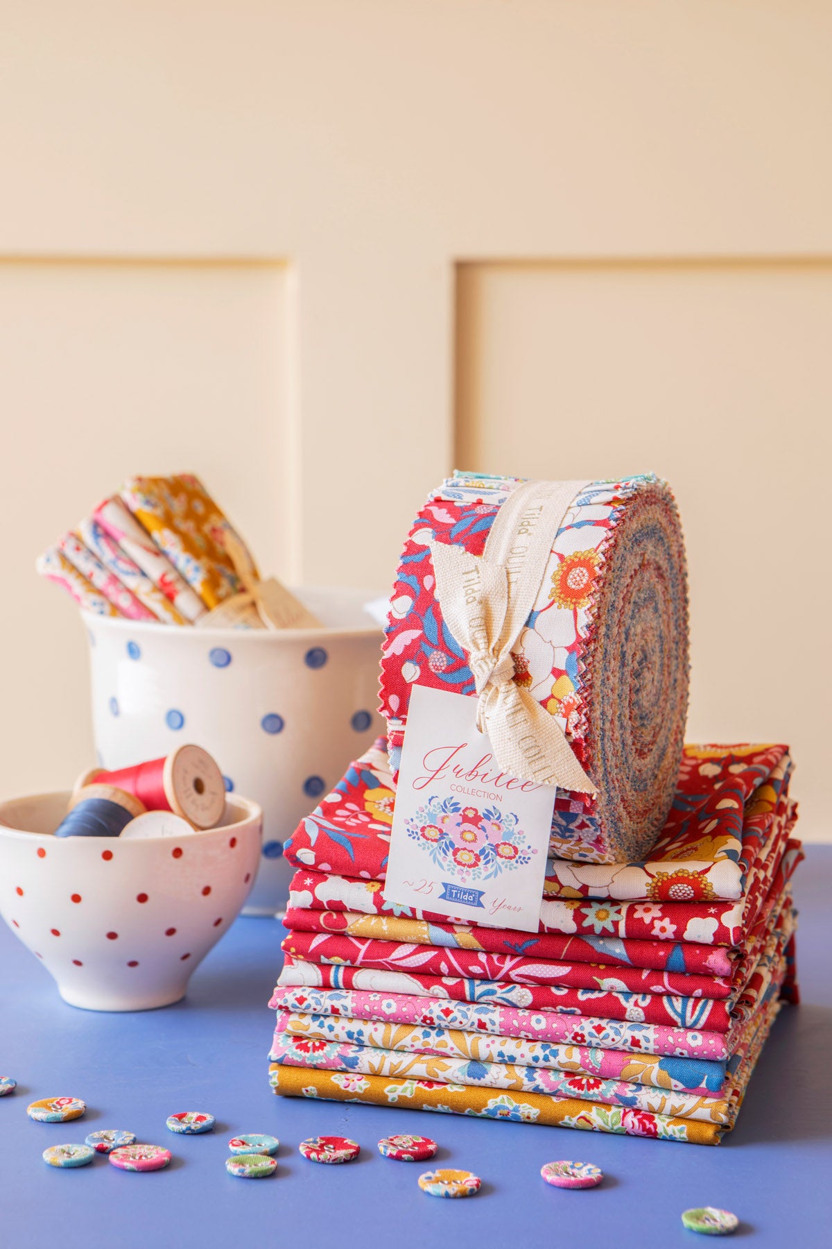 A stack of colorful fabric bundles and a rolled piece of patterned fabric tied with a cream ribbon and tag, placed on a blue surface against a warm beige background. A white polka-dot cup and a small white bowl with red dots hold additional spools and fabric pieces.
Photo by Tildafabrics.com [additional_image_link]