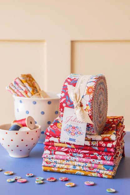 A stack of colorful fabric bundles and a rolled piece of patterned fabric tied with a cream ribbon and tag, placed on a blue surface against a warm beige background. A white polka-dot cup and a small white bowl with red dots hold additional spools and fabric pieces.
Photo by Tildafabrics.com [additional_image_link]