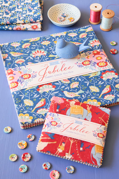 Two fabric stacks from the Jubilee collection, one blue with floral and bird patterns and one red with elephant and floral patterns, displayed on a lavender surface with spools of thread, buttons, and a small dish.
Photo by Tildafabrics.com [additional_image_link]