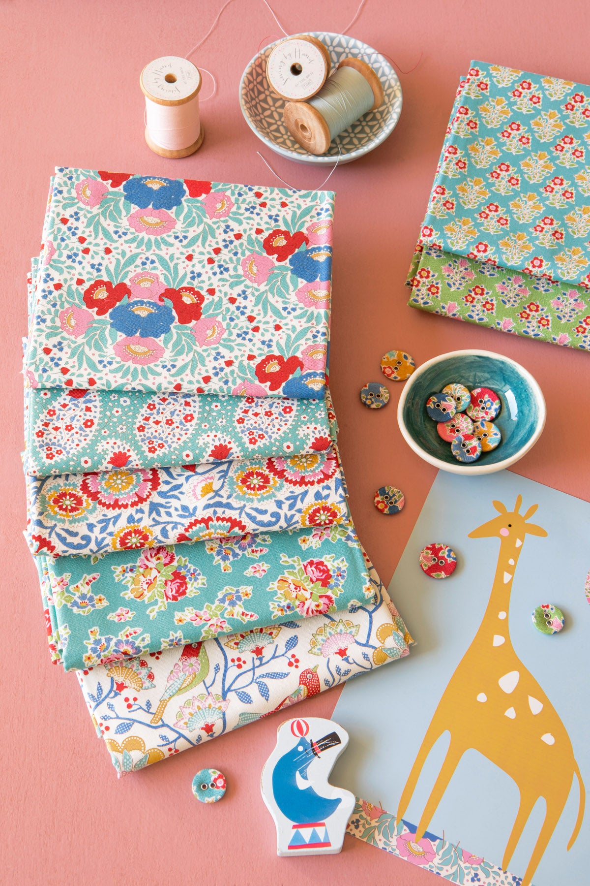 A styled flat-lay of folded fabric samples with colorful floral and paisley patterns, arranged on a pink background with spools of thread, buttons, a giraffe cutout, and small decorative pieces.
Photo by Tildafabrics.com [additional_image_link]