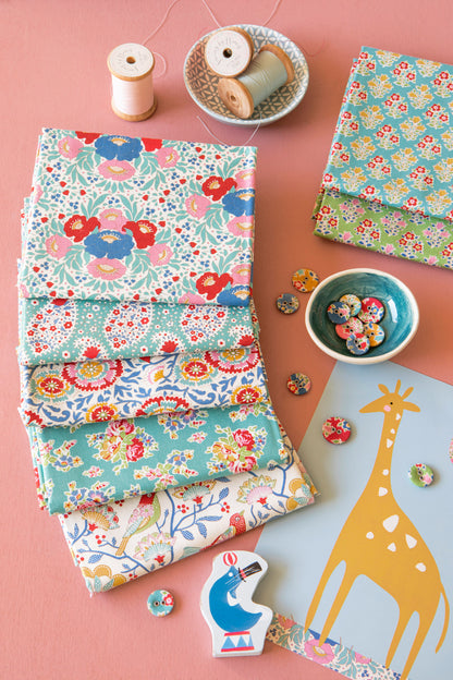 A styled flat-lay of folded fabric samples with colorful floral and paisley patterns, arranged on a pink background with spools of thread, buttons, a giraffe cutout, and small decorative pieces.
Photo by Tildafabrics.com [additional_image_link]