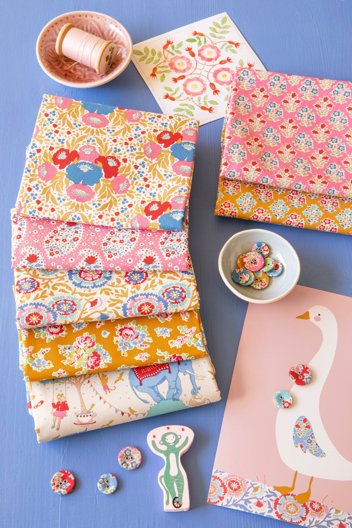 A styled flat-lay of fabric bolts with colorful floral and whimsical patterns, alongside sewing buttons, a spool of thread, and a printed card featuring a white goose on a pink background, all arranged on a blue surface.
Photo by Tildafabrics.com [additional_image_link]
