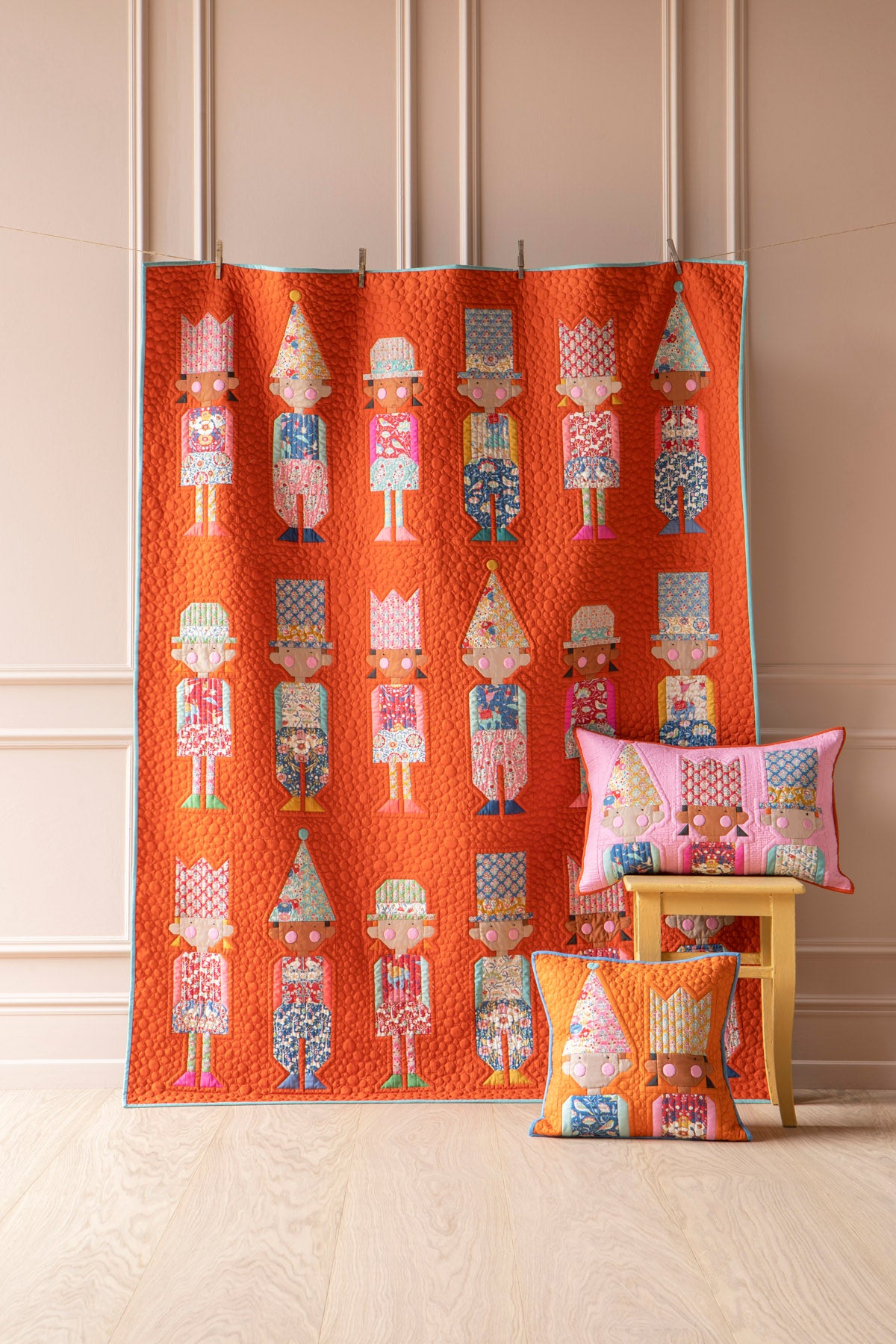 An orange quilted fabric panel featuring a repeating pattern of colorful nutcracker figures, displayed hanging on a stand in a room with beige paneled walls and light wood flooring. Two matching decorative pillows with the same nutcracker pattern are styled on a small wooden stool.
Photo by Tildafabrics.com [image_link]