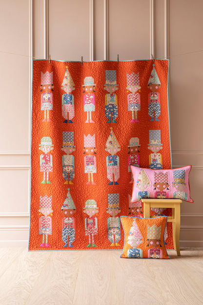 An orange quilted fabric panel featuring a repeating pattern of colorful nutcracker figures, displayed hanging on a stand in a room with beige paneled walls and light wood flooring. Two matching decorative pillows with the same nutcracker pattern are styled on a small wooden stool.
Photo by Tildafabrics.com [image_link]