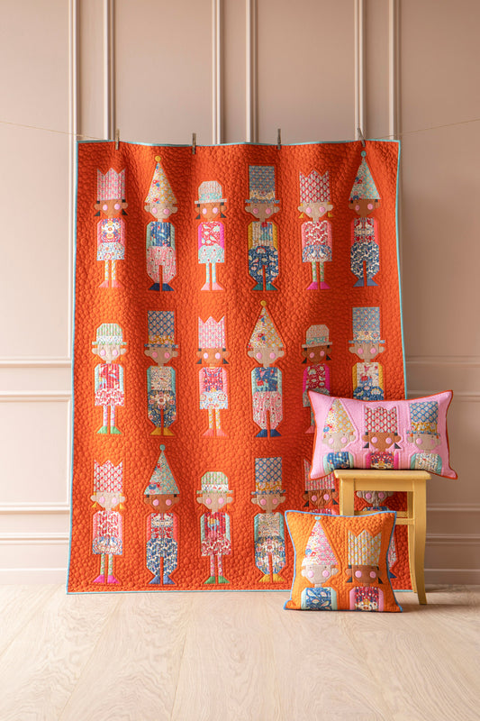 An orange quilted fabric panel featuring a repeating pattern of colorful nutcracker figures, displayed hanging on a stand in a room with beige paneled walls and light wood flooring. Two matching decorative pillows with the same nutcracker pattern are styled on a small wooden stool.
Photo by Tildafabrics.com [image_link]