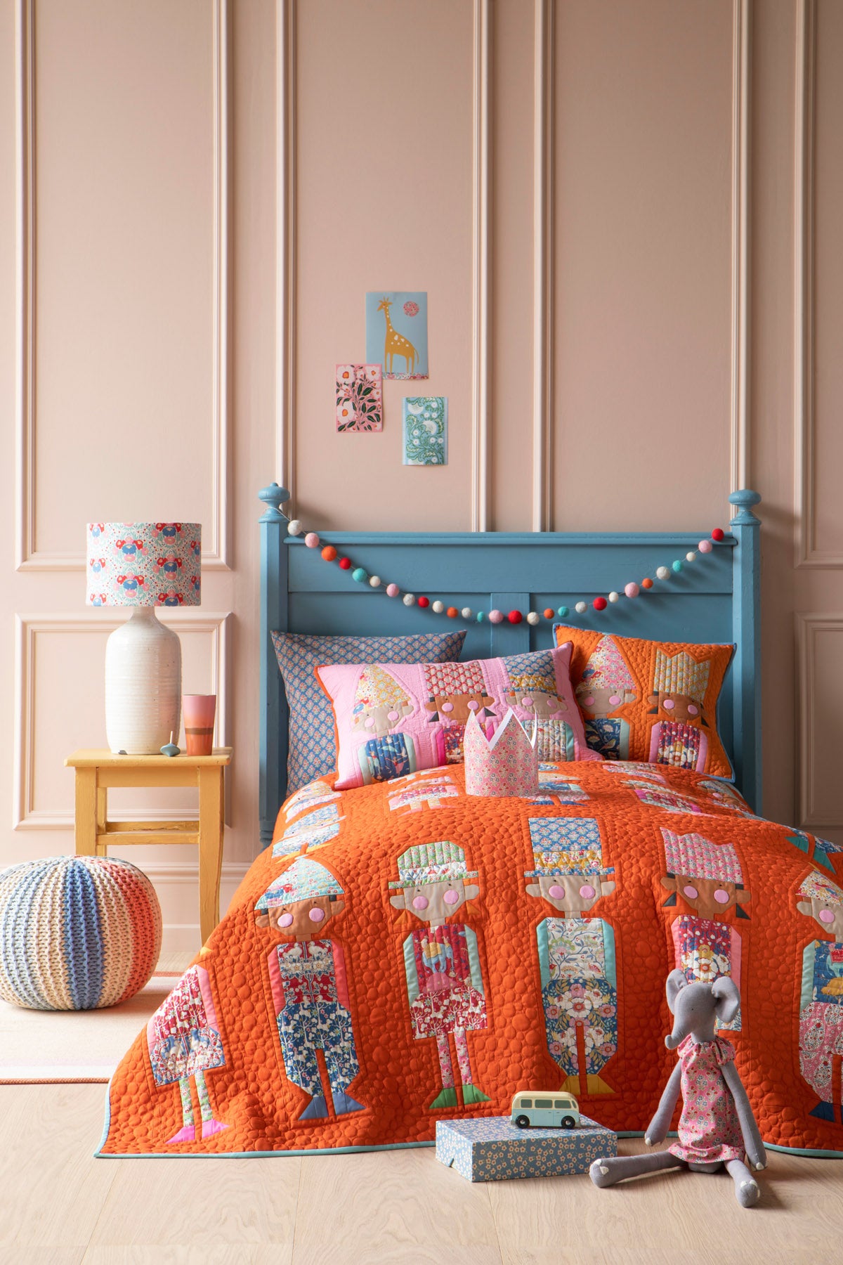 A children's bed styled in a nursery setting, featuring a bright orange quilted bedspread and matching pillow shams with a whimsical character print. The bed has a light blue wooden headboard decorated with a colorful bunting garland, set against a pink paneled wall with a small wooden side table and lamp.
Photo by Tildafabrics.com [additional_image_link]