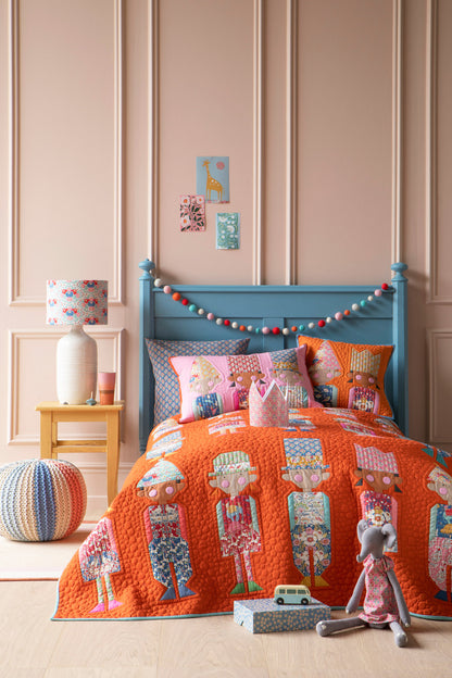A children's bed styled in a nursery setting, featuring a bright orange quilted bedspread and matching pillow shams with a whimsical character print. The bed has a light blue wooden headboard decorated with a colorful bunting garland, set against a pink paneled wall with a small wooden side table and lamp.
Photo by Tildafabrics.com [additional_image_link]