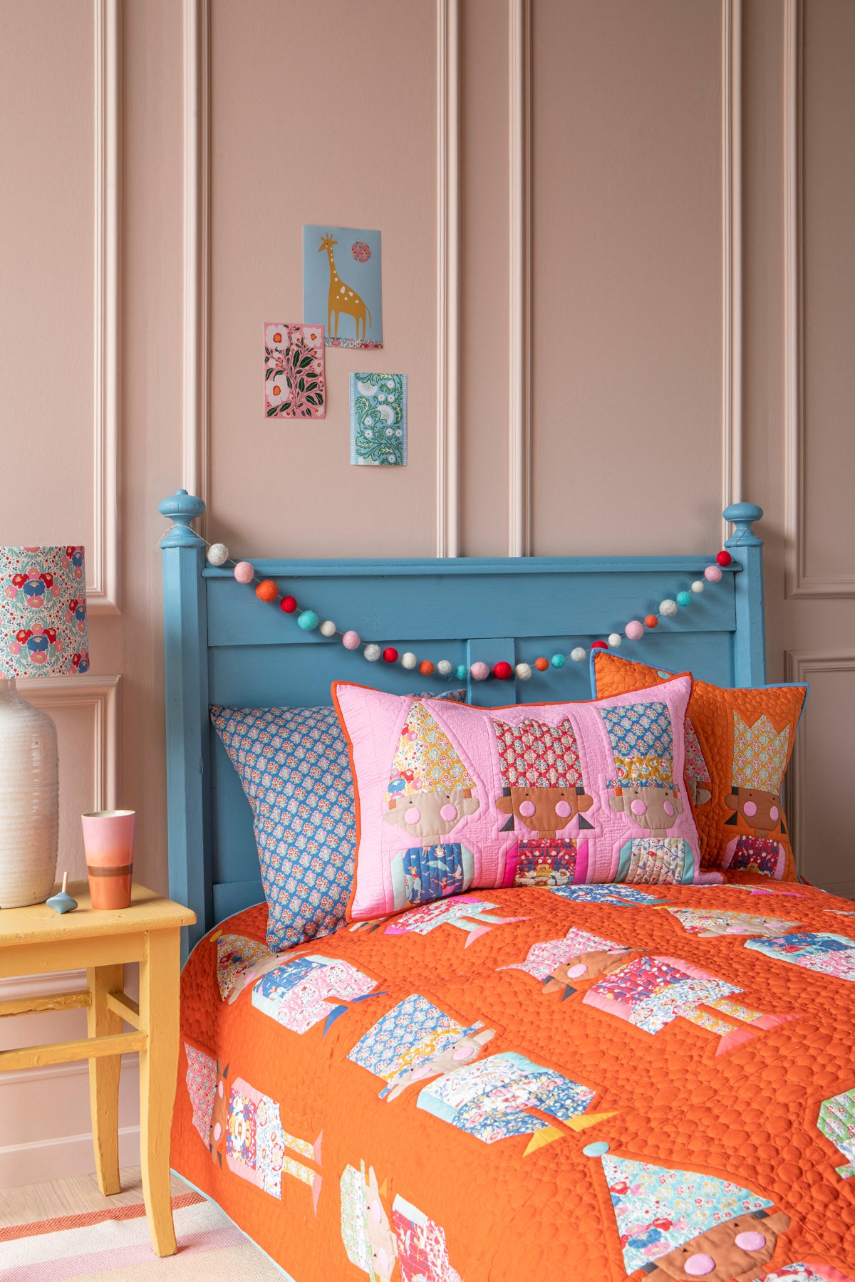 A styled children's bedroom scene showing a bed with a bright blue wooden headboard, orange quilted bedding, patterned pillows, and a colorful beaded garland against paneled pink walls.
Photo by Tildafabrics.com [additional_image_link]