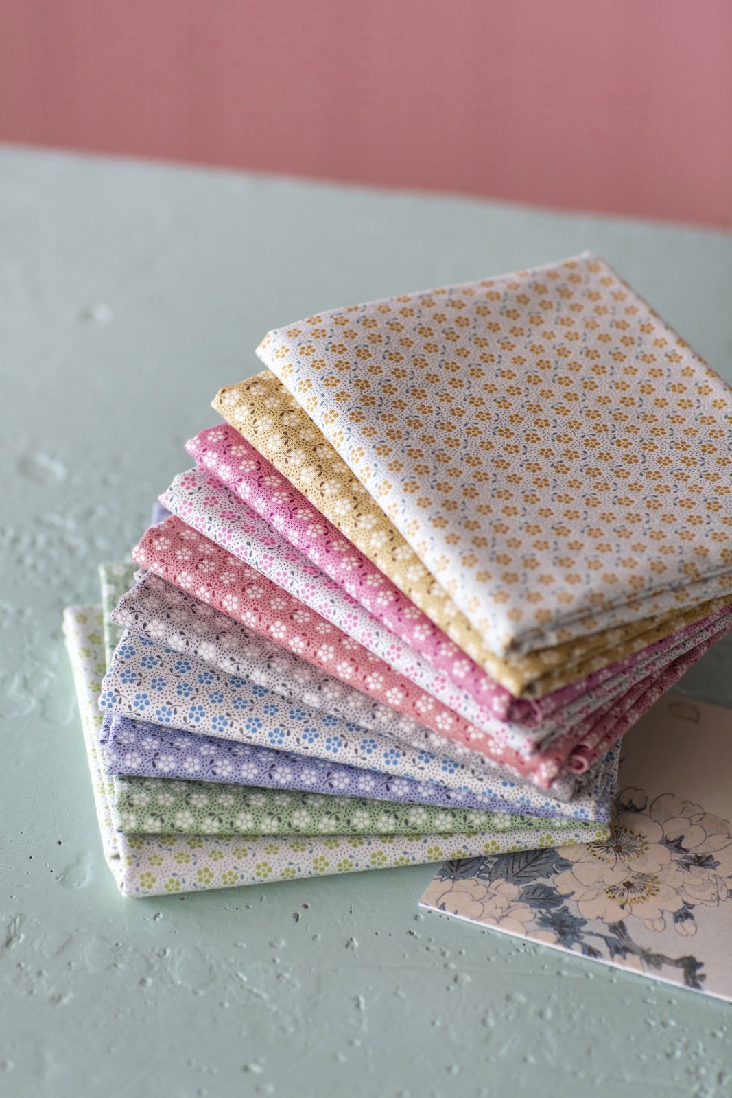 A stack of folded patterned fabric squares in soft pastel colors, including cream, yellow, pink, purple, and green, arranged on a light gray surface against a pink background.
Photo by Tildafabrics.com [additional_image_link]