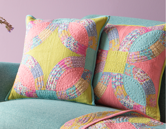 A styled interior scene with a pastel lavender wall, framed art prints, a mint green sofa, two patterned pink and yellow throw pillows, and a matching patterned quilt draped over the arm. A round pink side table with a vase of flowers and small bowls sits beside the sofa.
Photo by Tildafabrics.com [image_link]