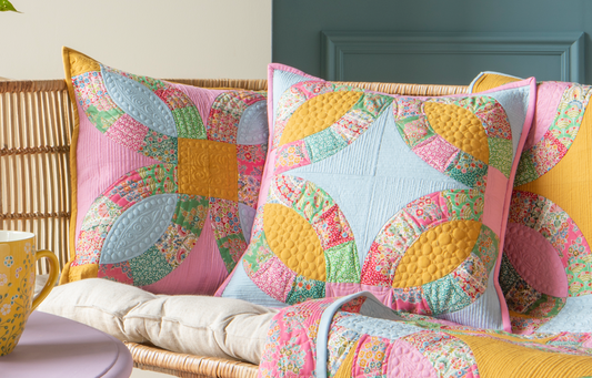 A styled interior scene showing a pastel quilted throw blanket and matching decorative pillows with a colorful butterfly patchwork pattern, arranged on a sofa in a room with blue paneled walls and a lavender side table.
Photo by Tildafabrics.com [image_link]