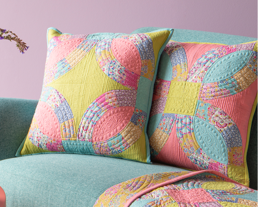 A styled interior scene with a pastel lavender wall, framed art prints, a mint green sofa, two patterned pink and yellow throw pillows, and a matching patterned quilt draped over the arm. A round pink side table with a vase of flowers and small bowls sits beside the sofa.
Photo by Tildafabrics.com [image_link]