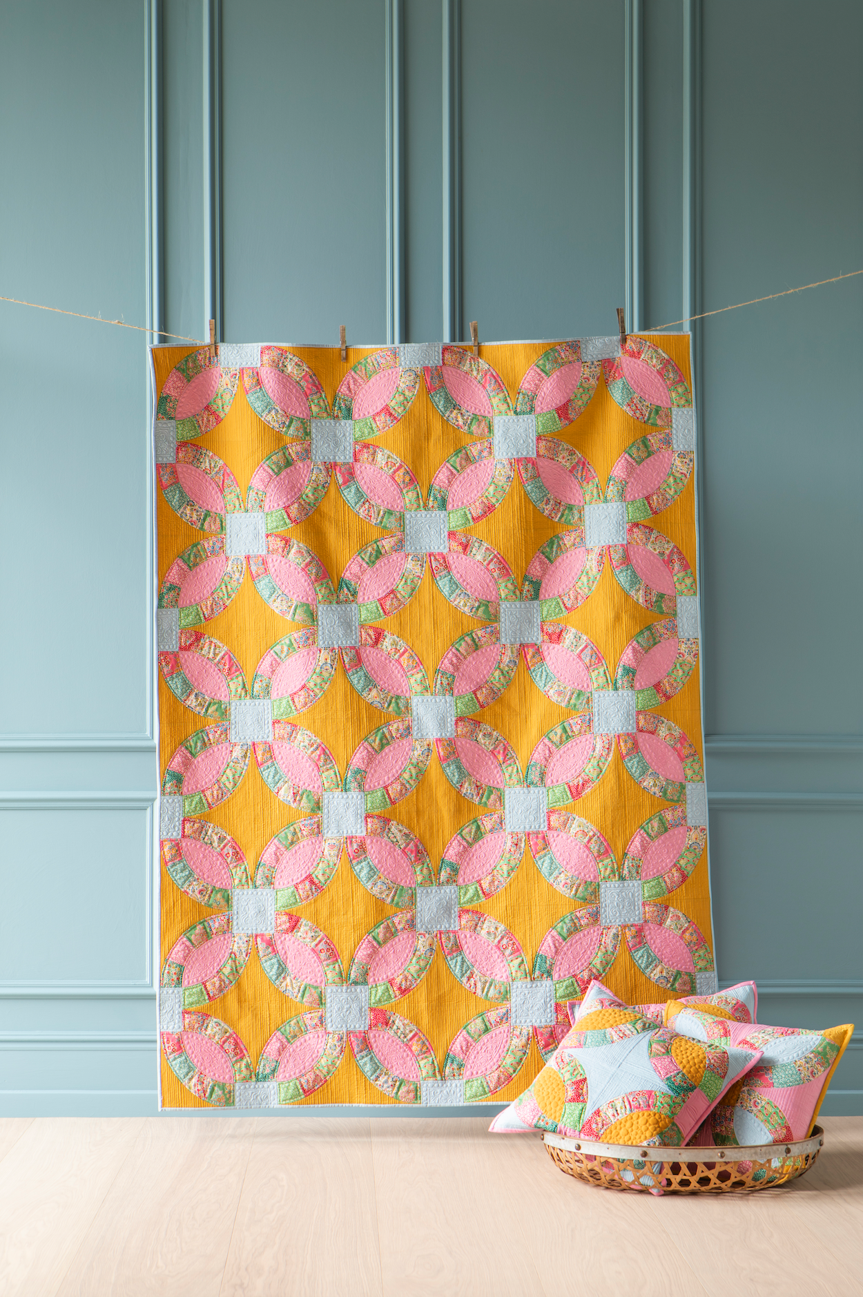 A quilt with a repeating pink and white wedding ring pattern on a mustard yellow background, hanging from clothespins against a blue paneled wall. Two matching decorative pillows rest in a woven basket on the floor below.
Photo by Tildafabrics.com [additional_image_link]