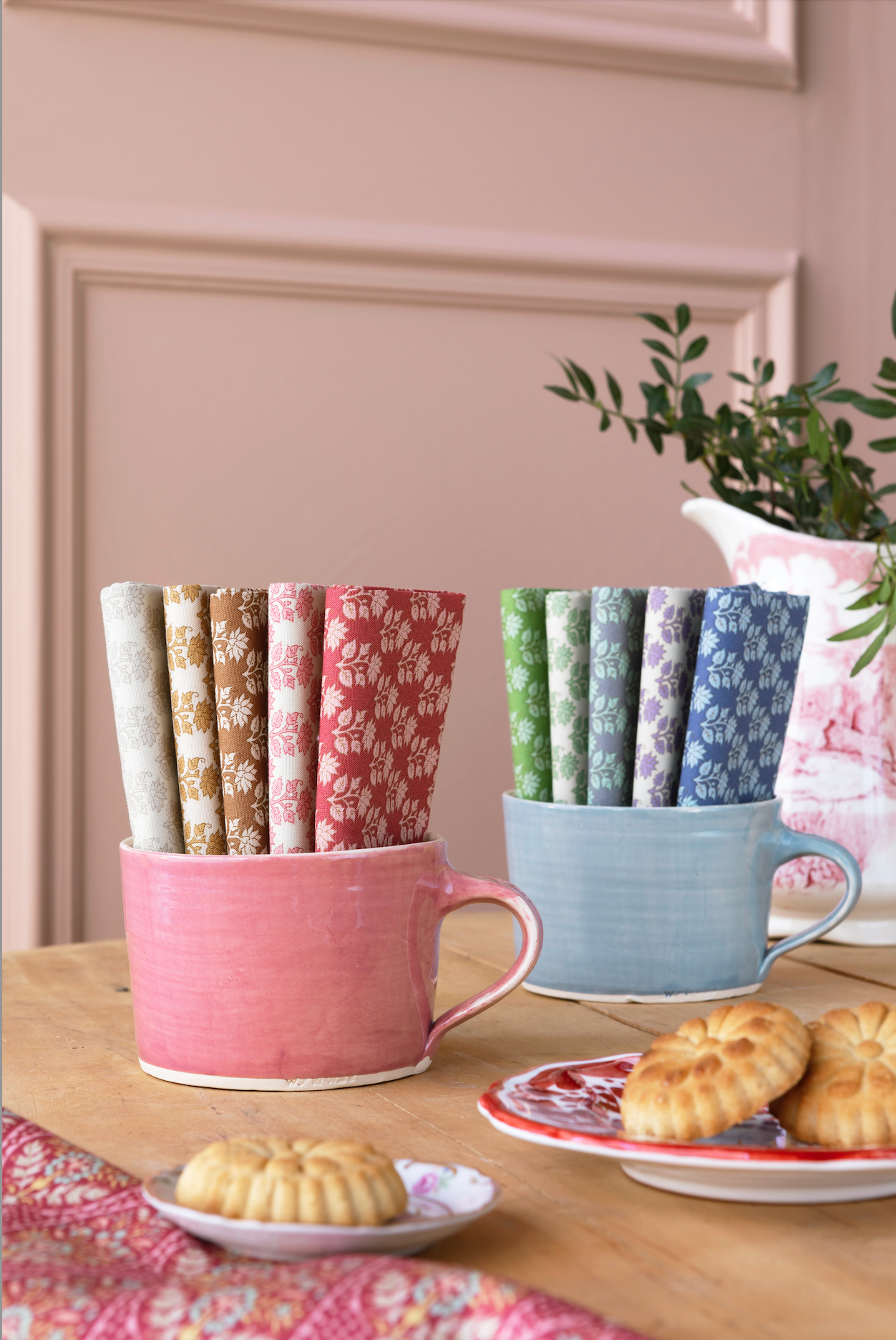 Two ceramic mugs holding multiple rolled fabric napkins with different floral patterns, styled on a wooden table with cookies and a potted plant in the background.
Photo by Tildafabrics.com [additional_image_link]