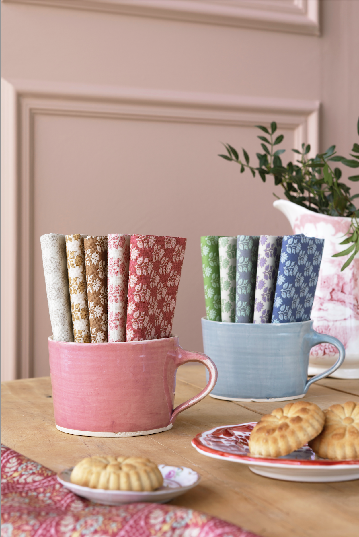 Two ceramic mugs holding multiple rolled fabric napkins with different floral patterns, styled on a wooden table with cookies and a potted plant in the background.
Photo by Tildafabrics.com [additional_image_link]