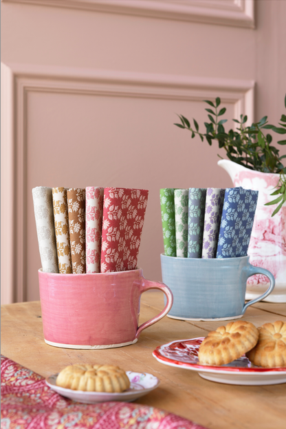 Two ceramic mugs holding multiple rolled fabric napkins with different floral patterns, styled on a wooden table with cookies and a potted plant in the background.
Photo by Tildafabrics.com [additional_image_link]