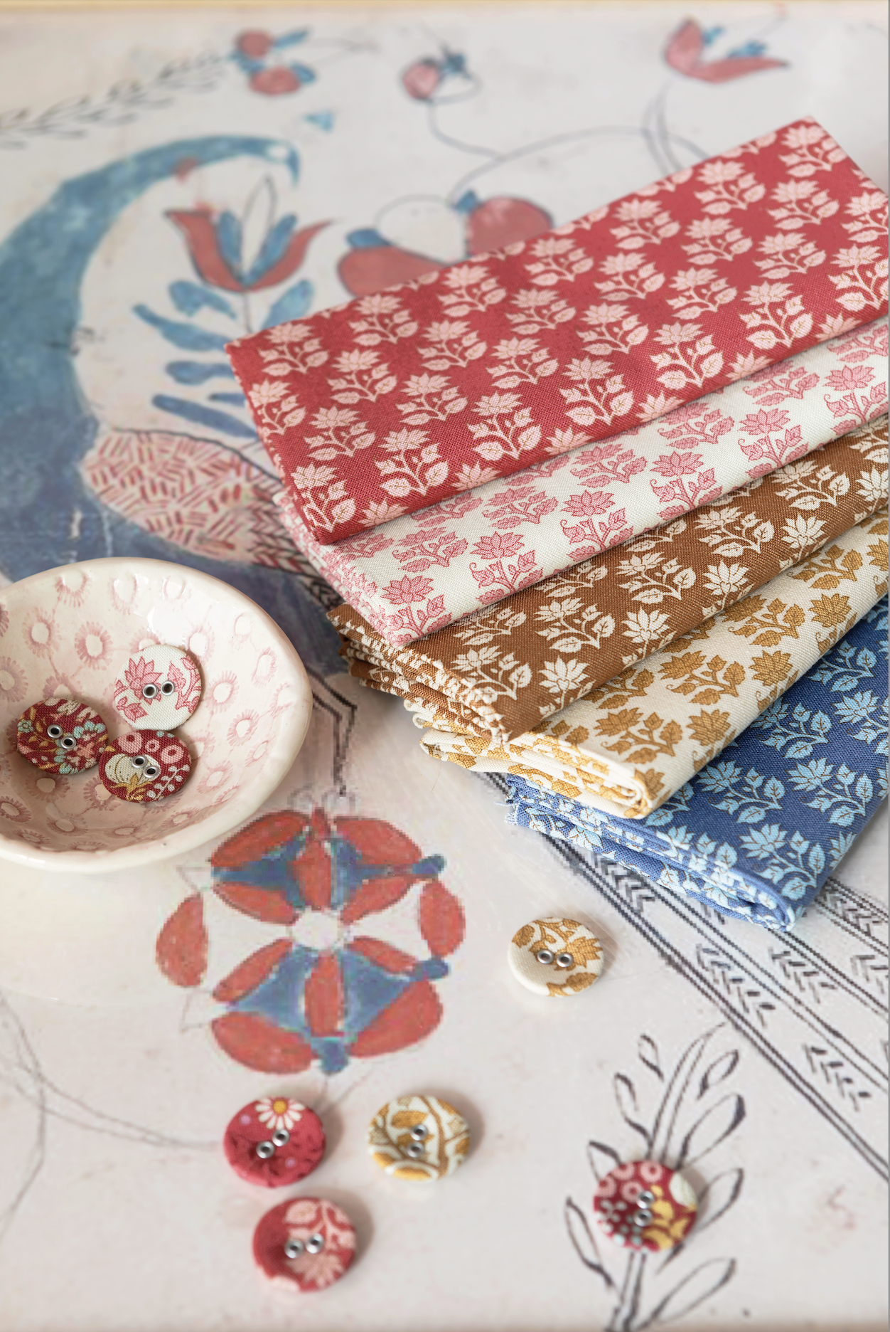A stack of four patterned fabric bolts in red, tan, and blue floral prints, styled on a decorative printed paper background with small buttons.
Photo by Tildafabrics.com [additional_image_link]