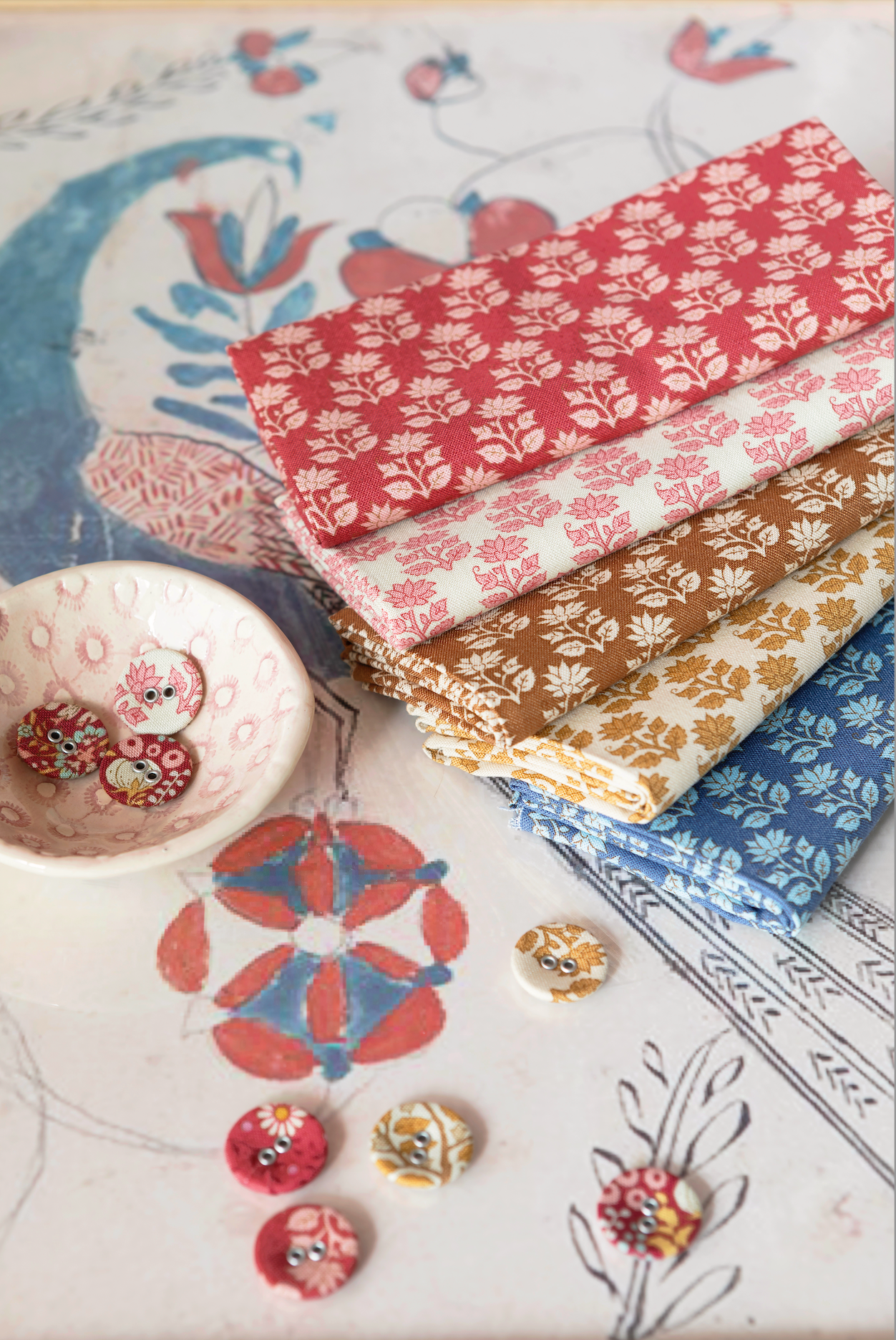 A stack of four patterned fabric bolts in red, tan, and blue floral prints, styled on a decorative printed paper background with small buttons.
Photo by Tildafabrics.com [additional_image_link]