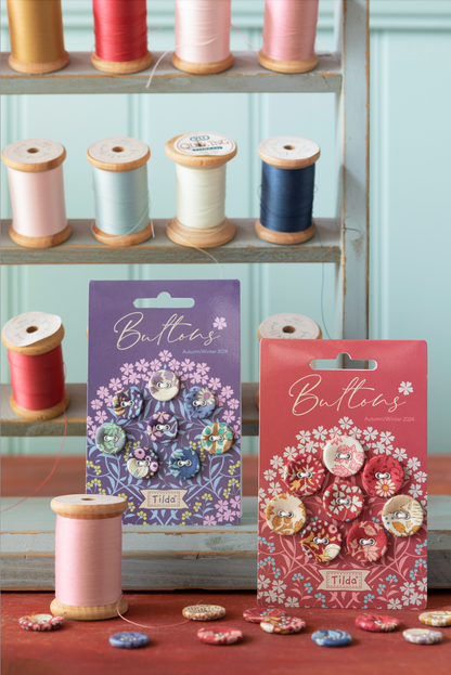 Two packaged buttons labeled 'Buttons' and 'Tilda' displayed in front of shelves of spools of thread. The left package is blue and the right package is red, both featuring decorative button illustrations.
Photo by Tildafabrics.com [additional_image_link]