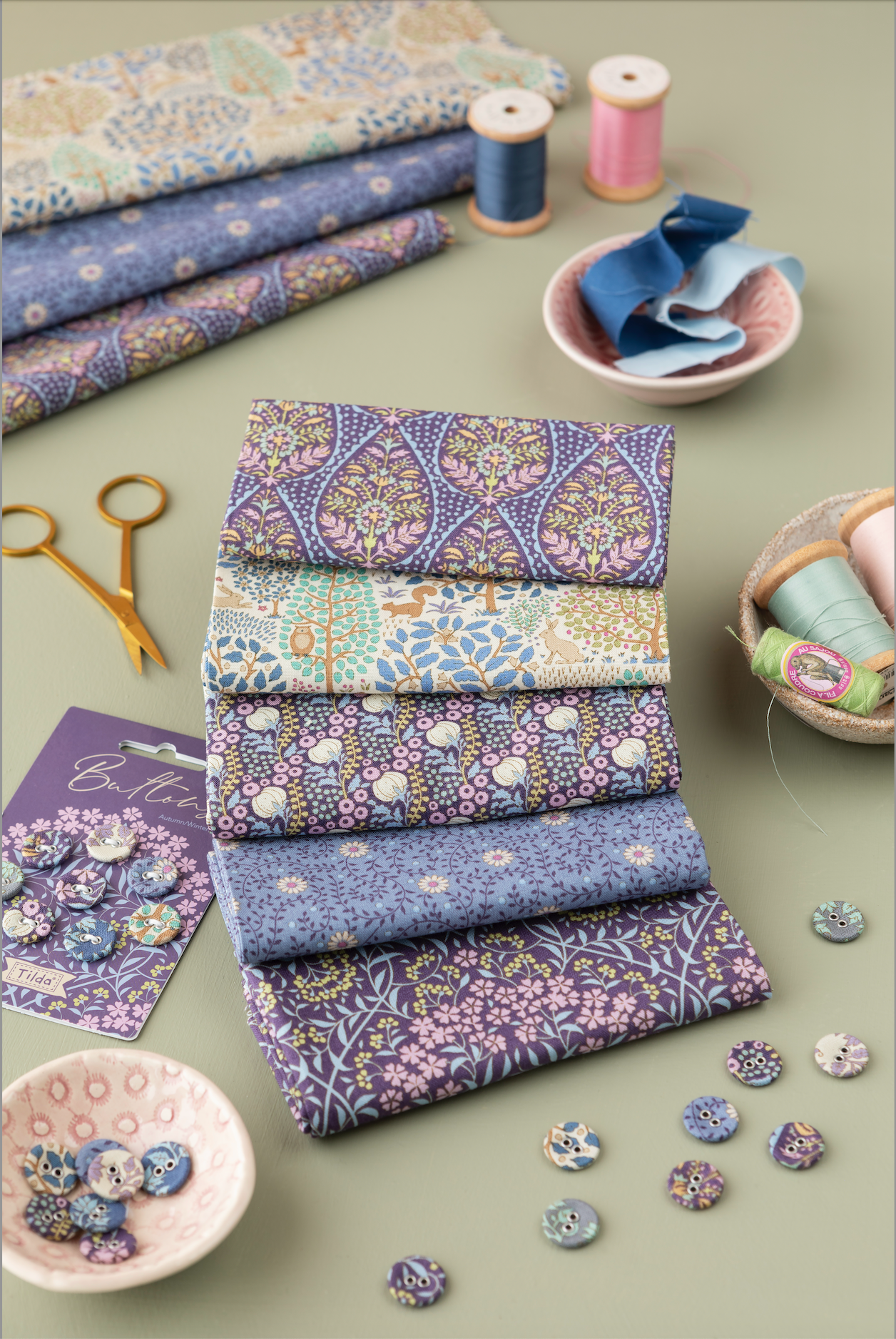 A styled flat-lay of folded patterned fabrics in soft blue, lavender, beige, and cream tones, arranged with sewing props including spools of thread, scissors, buttons, and small bowls on a muted green background.
Photo by Tildafabrics.com [additional_image_link]