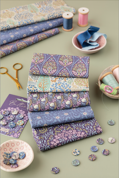 A styled flat-lay of folded patterned fabrics in soft blue, lavender, beige, and cream tones, arranged with sewing props including spools of thread, scissors, buttons, and small bowls on a muted green background.
Photo by Tildafabrics.com [additional_image_link]