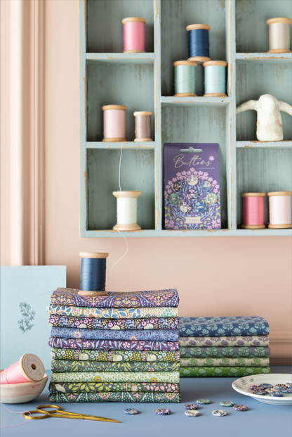 Stacks of patterned fabric bolts and spools of thread arranged on a table in front of a light blue shelving unit, with a purple fabric box labeled 'Boules' in the background.
Photo by Tildafabrics.com [additional_image_link]