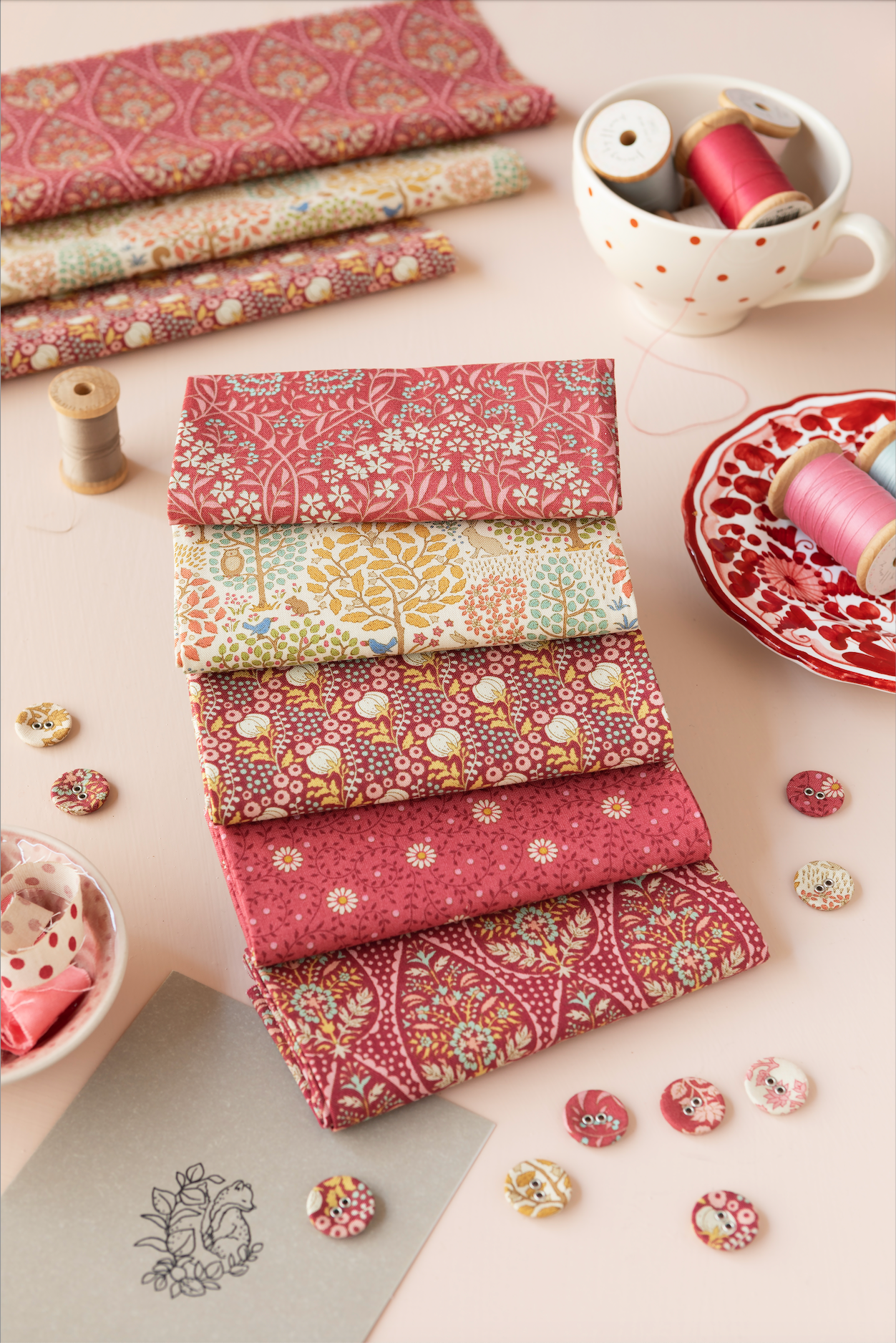 A styled flat-lay of a fabric collection set featuring six folded fabric squares with various floral and paisley patterns in dusty pink, sage green, and cream. The fabrics are arranged on a light beige surface alongside sewing props including thread spools in a white mug and a plate, decorative buttons, and a sketchbook.
Photo by Tildafabrics.com [additional_image_link]