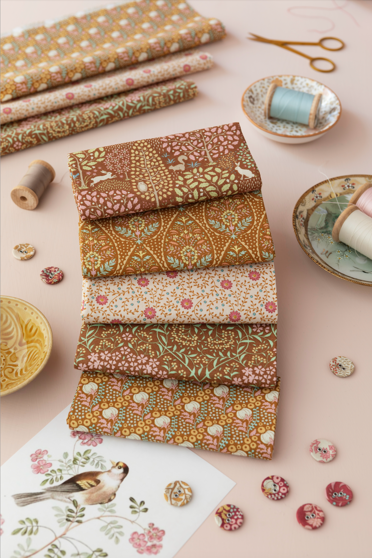 A styled flat-lay of six folded fabrics with intricate floral and bird patterns in earthy tones, arranged on a dusty pink background with sewing props like spools, buttons, and scissors.
Photo by Tildafabrics.com [additional_image_link]