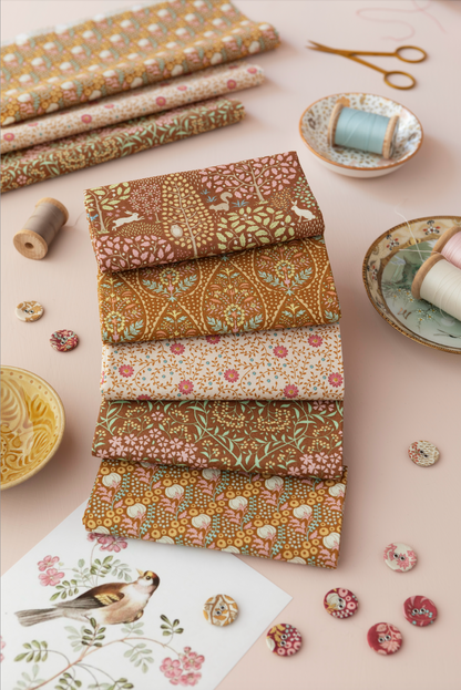 A styled flat-lay of six folded fabrics with intricate floral and bird patterns in earthy tones, arranged on a dusty pink background with sewing props like spools, buttons, and scissors.
Photo by Tildafabrics.com [additional_image_link]