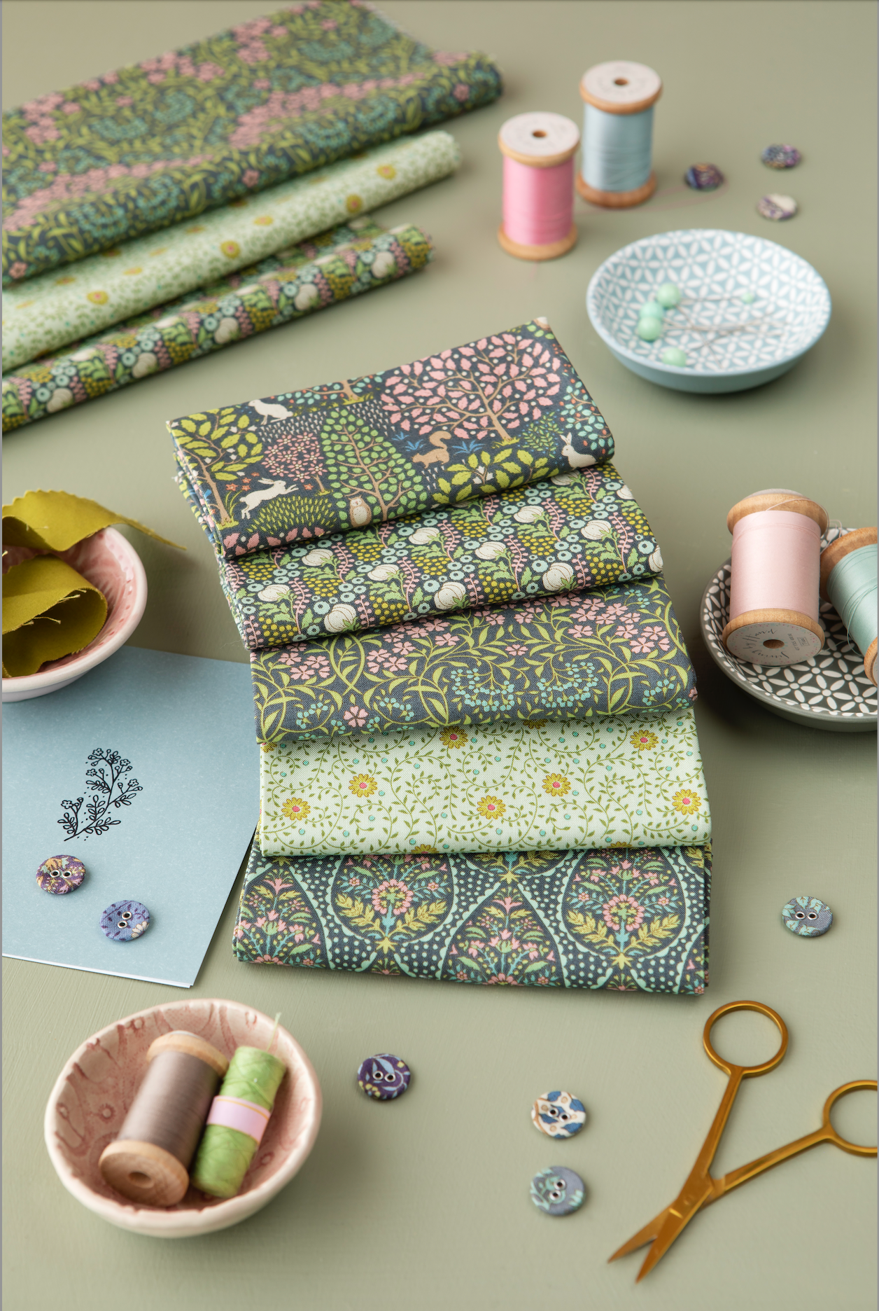 A styled flat-lay of multiple fabrics and spools of thread with assorted sewing accessories, including buttons, scissors, and small bowls, arranged on a neutral beige surface.
Photo by Tildafabrics.com [additional_image_link]