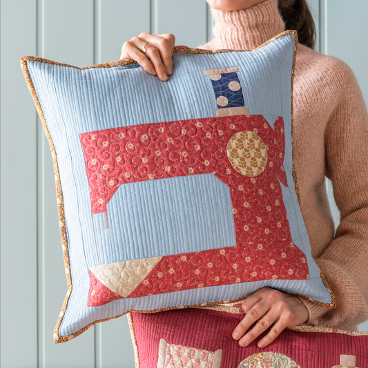 A person holding a quilted pillow with a blue chambray background and a sewing machine motif. The pillow has a Tilda brand label.
Photo by Tildafabrics.com [image_link]