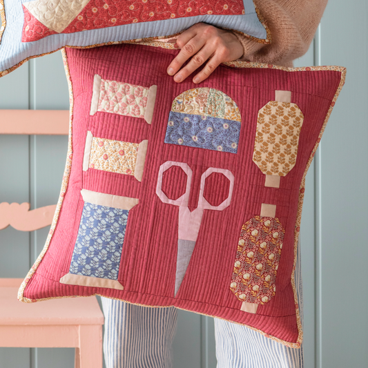 A woman holds one square patchwork pillows. The pillow is red background with scissors, spools, pincusion design and cream accents.
Photo by Tildafabrics.com [image_link]