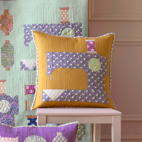 A completed pillow with a mustard yellow base and a variety of patches with sewing-themed designs, placed on a white wooden stand against a green textured background.
Photo by Tildafabrics.com [image_link]