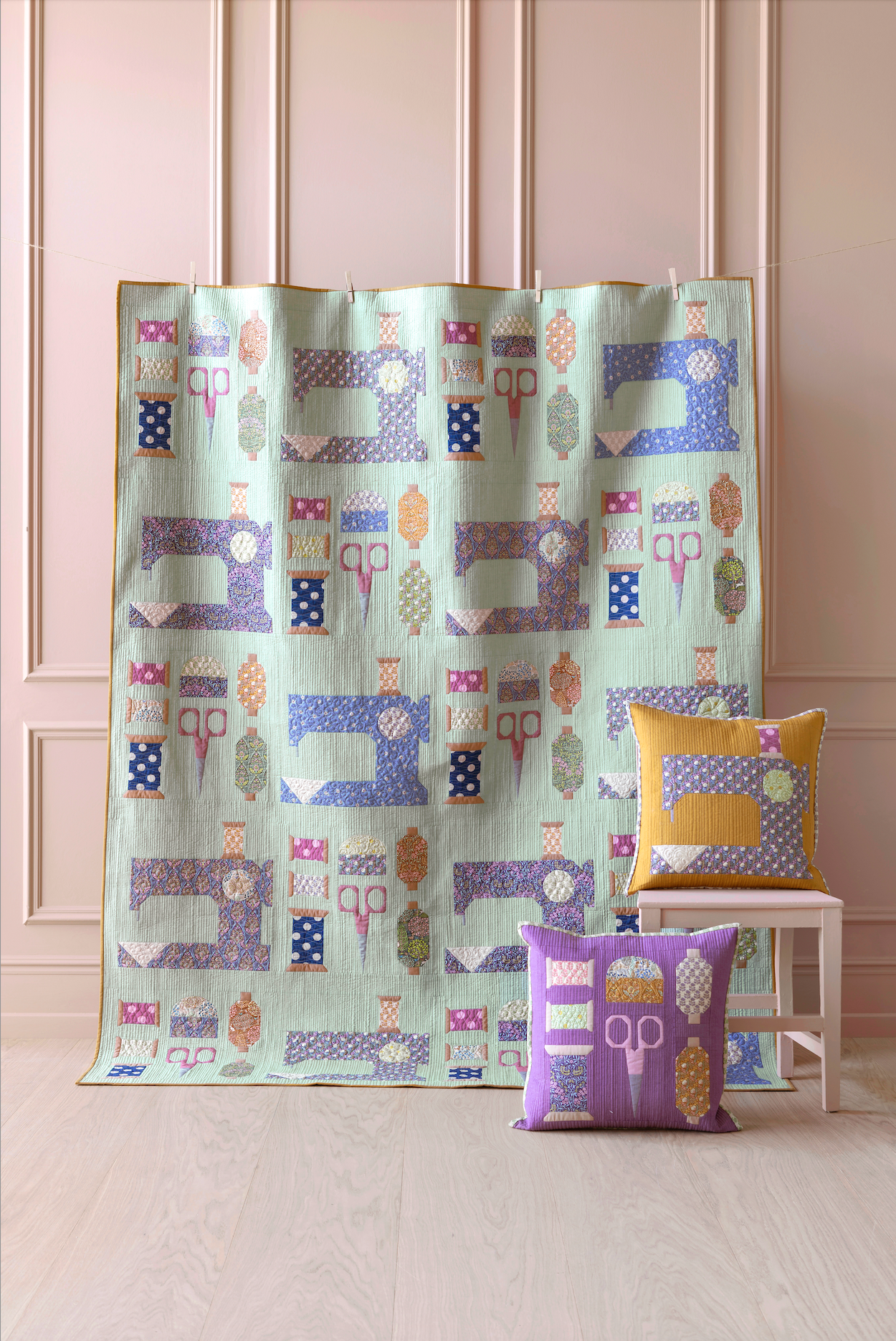 A quilted fabric panel or throw featuring a patchwork design of sewing machines and scissors in pastel colors, displayed on a stand in a room with a light wood floor and pink paneled wall. Two decorative pillows are styled on a small white stool next to it.
Photo by Tildafabrics.com [additional_image_link]
