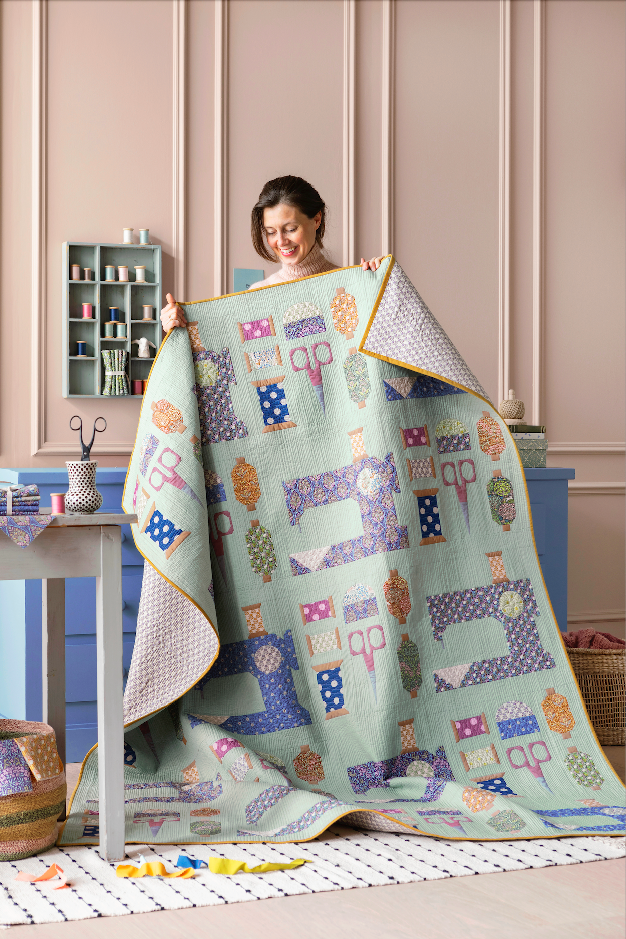 A woman holds up a large quilted blanket featuring a colorful patchwork design of sewing machines and quilting motifs on a pale mint green background. The scene is set in a cozy, styled sewing room with pastel pink paneled walls and blue cabinets.
Photo by Tildafabrics.com [additional_image_link]