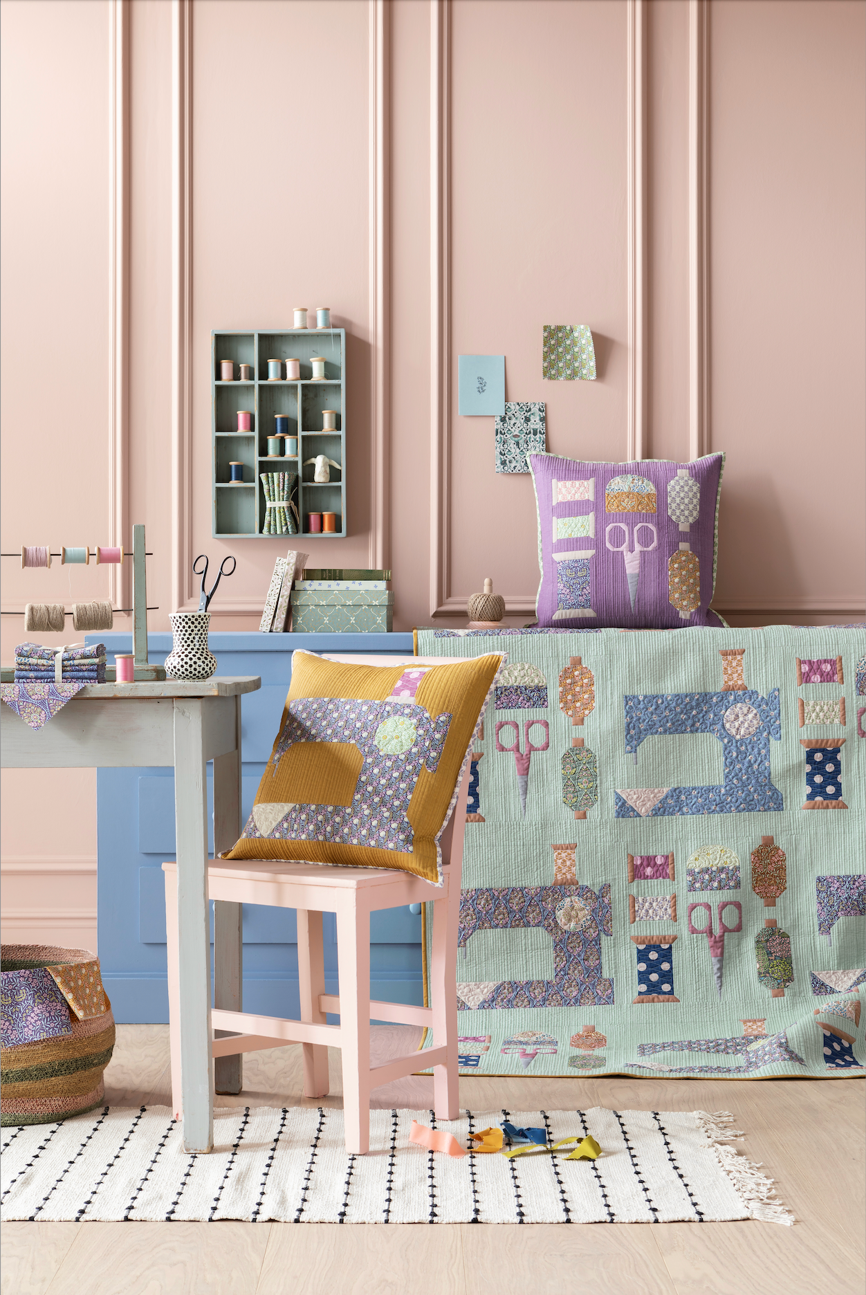A styled interior scene showing a patchwork quilt draped over a bench, a matching patterned throw pillow, and another pillow on a chair, in a room with pink paneled walls and a sewing table setup.
Photo by Tildafabrics.com [additional_image_link]