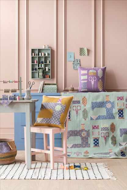 A styled interior scene showing a patchwork quilt draped over a bench, a matching patterned throw pillow, and another pillow on a chair, in a room with pink paneled walls and a sewing table setup.
Photo by Tildafabrics.com [additional_image_link]