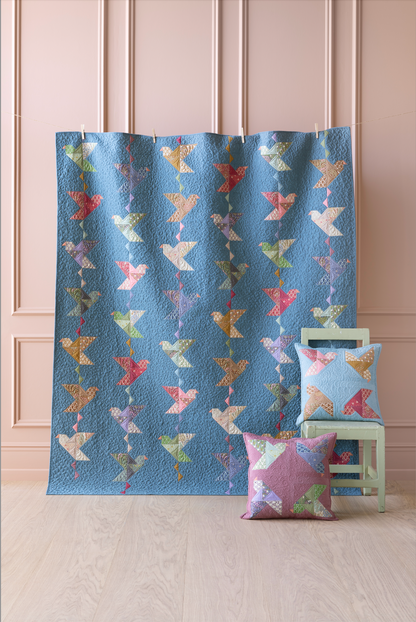 A blue quilted fabric panel with a repeating pattern of colorful birds in pastel shades, displayed in a room setting next to matching decorative pillows on a small chair.
Photo by Tildafabrics.com [additional_image_link]