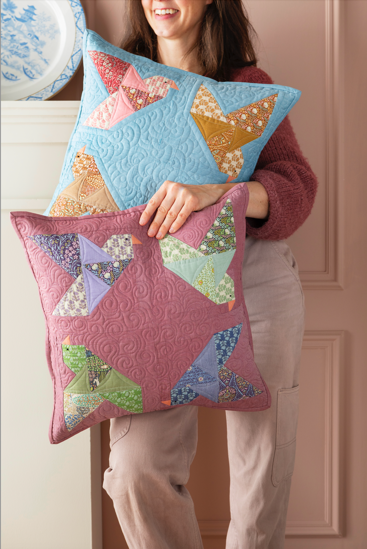 A woman holds two quilted square pillows with patchwork bird designs. The top pillow is light blue, and the bottom pillow is dusty pink.
Photo by Tildafabrics.com [additional_image_link]