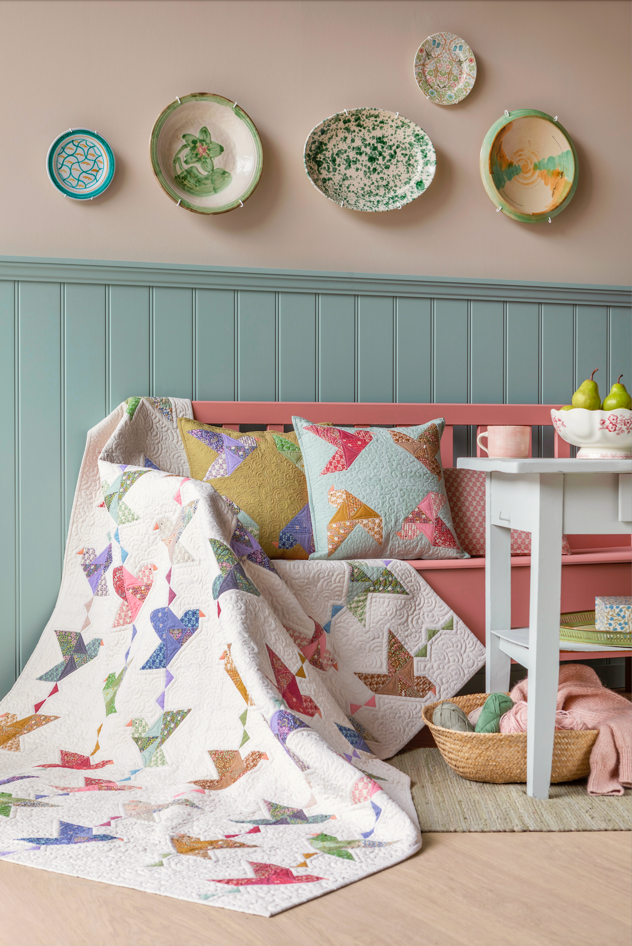 A cozy nursery or children's room scene with a quilted patchwork blanket draped over a pink bench, matching patterned pillows, a white side table, wall decor, and a soft rug on a light wood floor.
Photo by Tildafabrics.com [additional_image_link]