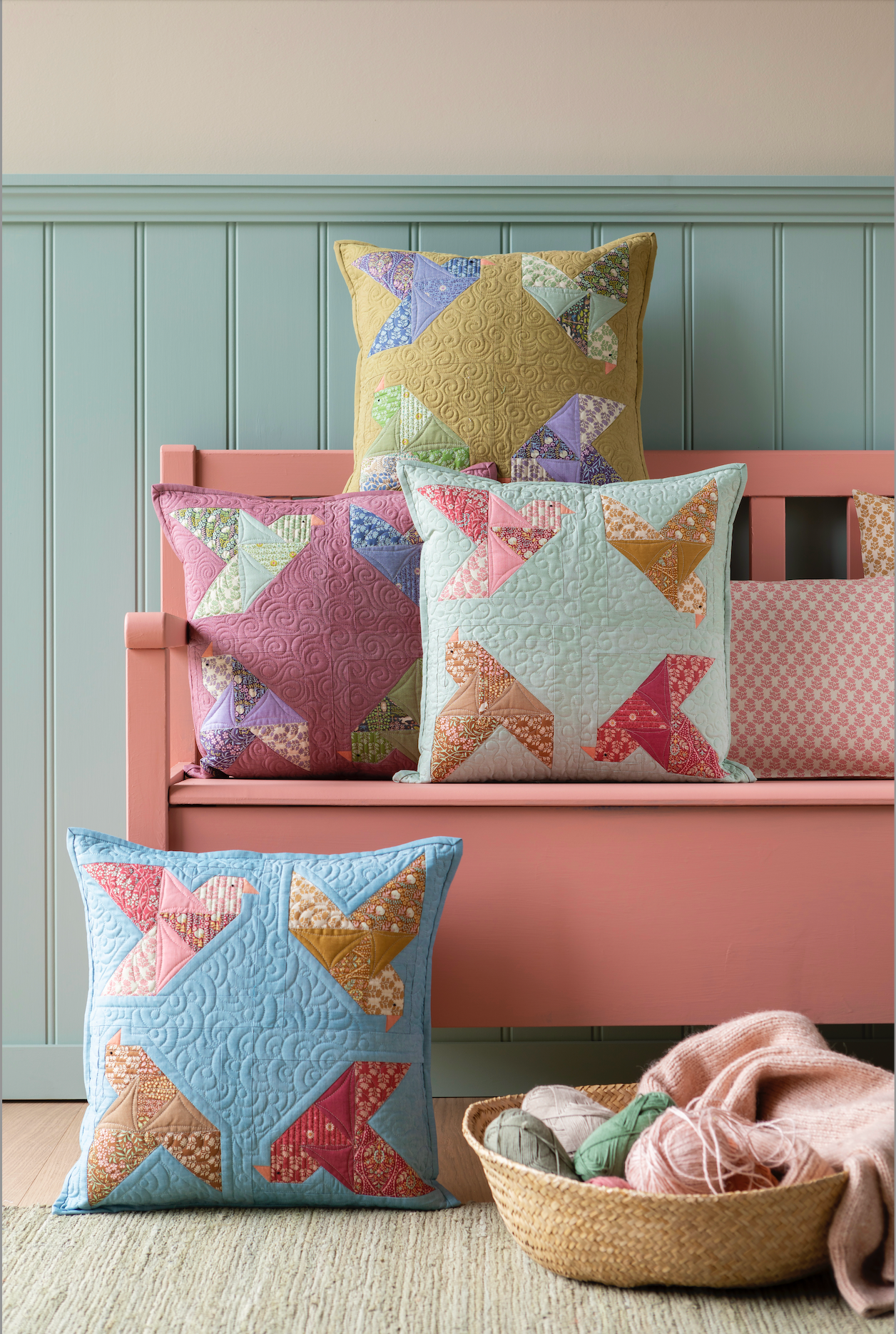 A set of colorful patchwork star-patterned quilted throw pillows styled on a pink wooden bench against a light blue paneled wall, with a matching pillow in the foreground and a basket of yarn and blanket on the floor.
Photo by Tildafabrics.com [additional_image_link]