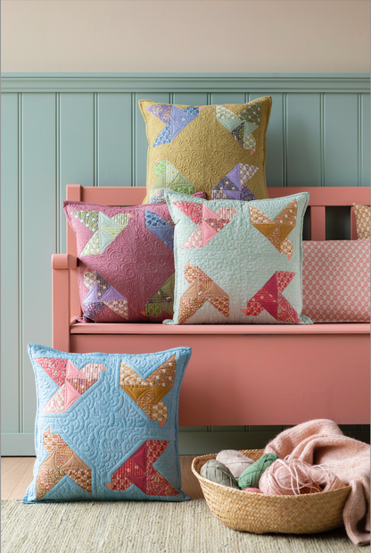 A set of colorful patchwork star-patterned quilted throw pillows styled on a pink wooden bench against a light blue paneled wall, with a matching pillow in the foreground and a basket of yarn and blanket on the floor.
Photo by Tildafabrics.com [additional_image_link]