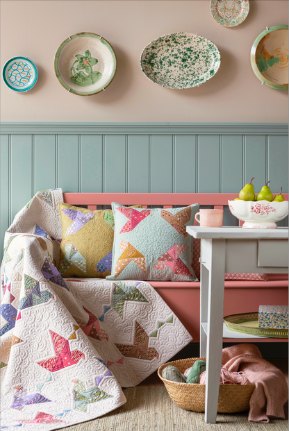 A styled interior scene showing a pink bench with colorful patchwork pillows and a matching quilt draped over it, alongside a white side table with fruit and a pink mug. Decorative ceramic plates hang on a beige wall above a blue paneled wainscoting.
Photo by Tildafabrics.com [additional_image_link]