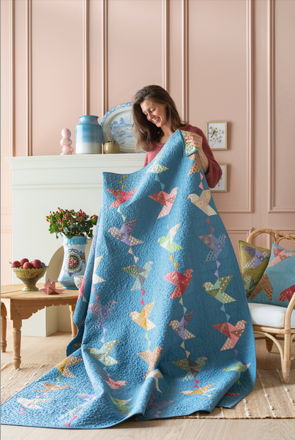 A woman holds up a large quilted blue blanket featuring a colorful pattern of paper cranes, set in a cozy room with pink paneled walls and a fireplace.
Photo by Tildafabrics.com [additional_image_link]
