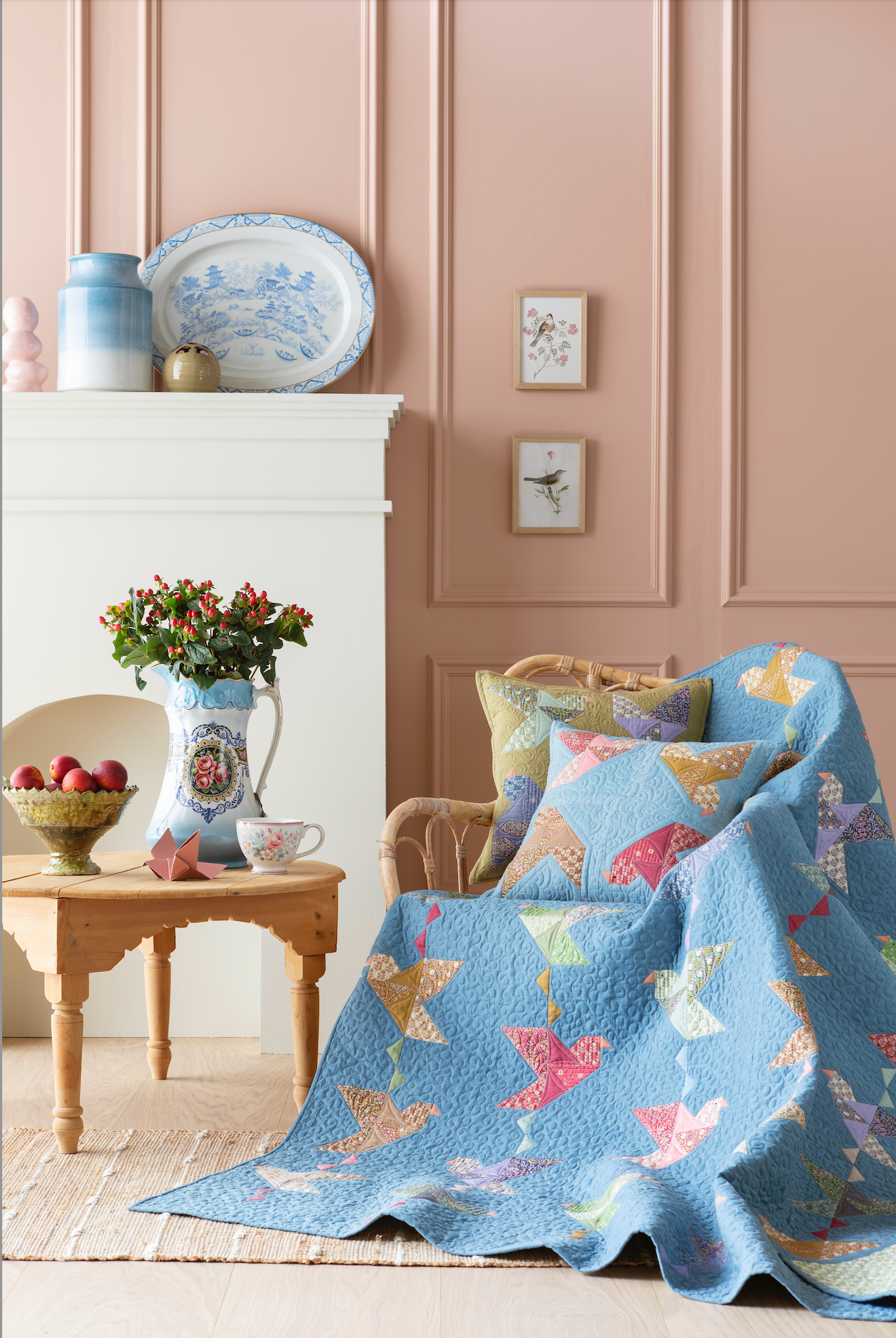 A light blue quilted throw blanket with a colorful butterfly pattern is draped over a wicker armchair in a styled room with pink paneled walls, a white fireplace, and a small wooden side table.
Photo by Tildafabrics.com [additional_image_link]