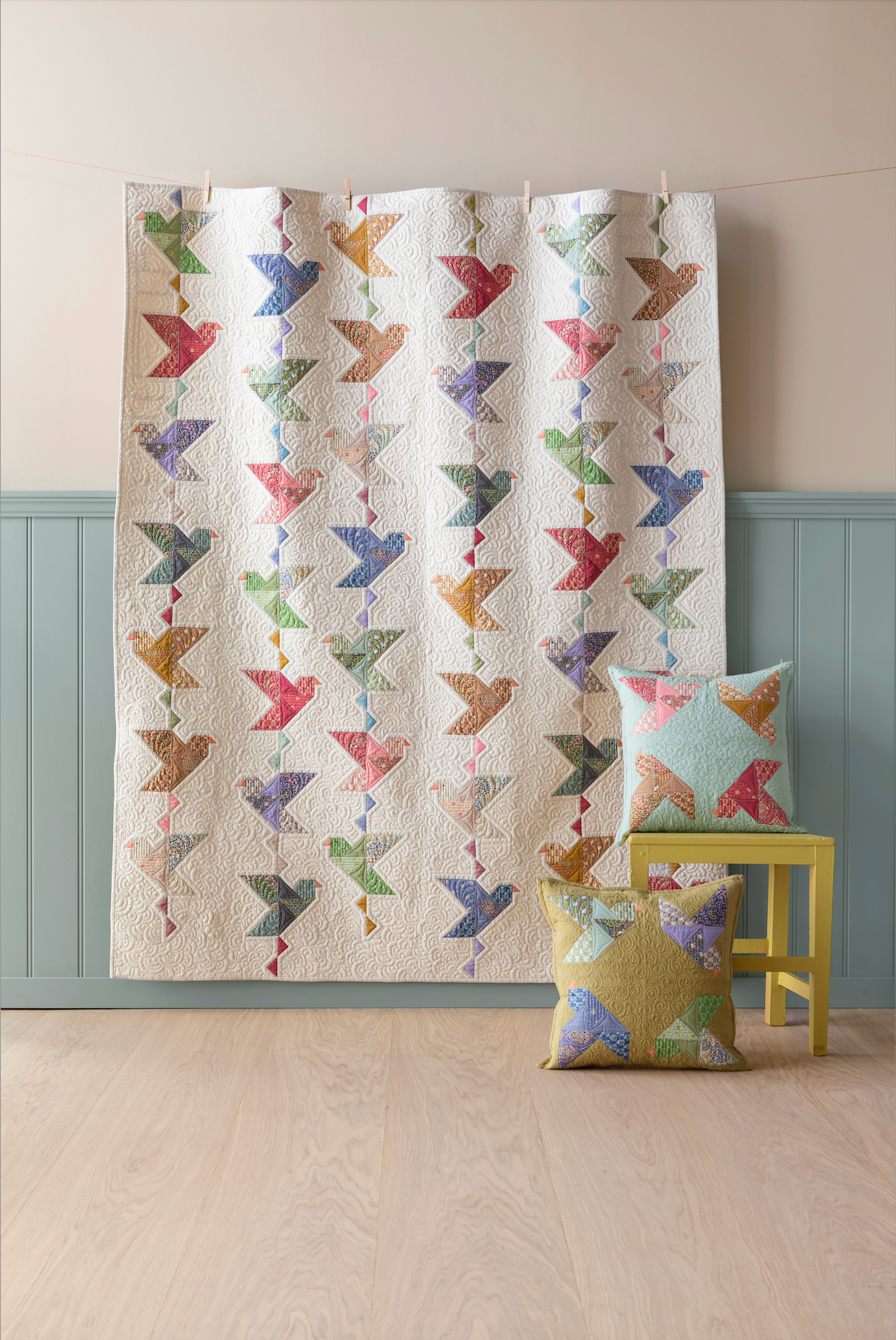 A quilted fabric panel or curtain featuring a repeating pattern of colorful birds on an off-white background, hanging in a room with a light wood floor, pale wall, and blue-green wainscoting. Two matching decorative pillows rest on a small yellow stool.
Photo by Tildafabrics.com [additional_image_link]
